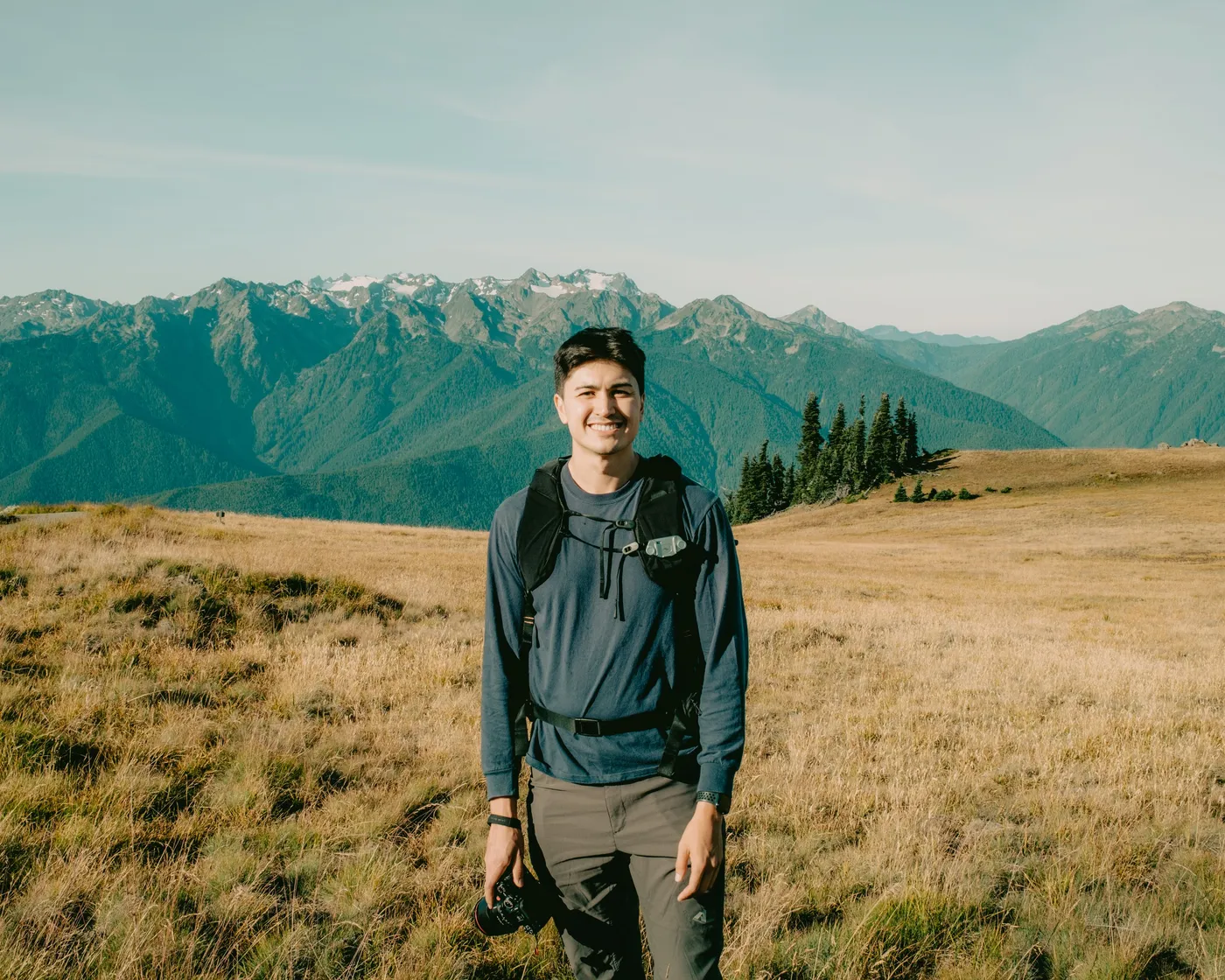 Omar Mussehl standing in a golden meadow below the Olympic mountains, camera in hand